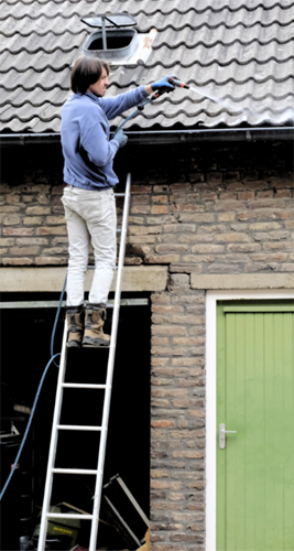 Foto van een persoon op een ladder voor het huis die de dakgoot schoon spuit